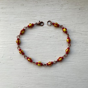 May include: A handmade bracelet with alternating red and yellow beads. The beads are strung on a copper chain and fastened with a copper clasp. The bracelet is shown on a white wooden surface.