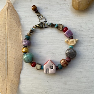 May include: A beaded bracelet featuring a central ceramic house charm, a small bird figurine, and various multicoloured beads in shades of red, blue, purple, and yellow. The bracelet includes a bronze-toned metal lobster clasp and an adjustable chain.