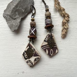 May include: Handmade earrings featuring a diamond-shaped pendant with a leaf charm. The earrings have a mix of brown, white, and bronze colors. The design includes wooden beads and a decorative pattern on the pendant.