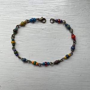 May include: A multi-coloured beaded bracelet with a bronze chain and clasp. The beads are small and come in various colours, including blue, green, yellow, orange, and red. The bracelet is displayed on a white wooden surface.