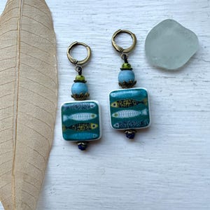 Peut inclure: Une paire de boucles d'oreilles pendantes avec des carreaux de céramique carrés sur fond sarcelle, ornés de petits poissons jaunes, blancs et bleus. Elles comportent des perles rondes bleu clair, des accents verts et des anneaux en métal bronze.