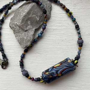May include: A handcrafted necklace featuring a central rectangular bead with a blue, red, and yellow pattern. The necklace is composed of various colorful beads in shades of black, purple, and blue, strung together with a metal clasp.