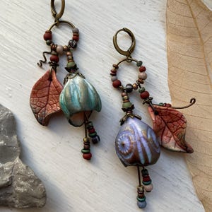 Peut inclure: Une paire de boucles d'oreilles pendantes avec des fermoirs dormeuses en bronze, des petites perles multicolores et des breloques en forme de feuilles texturées. L'une possède un pendentif cloche bleu-vert, l'autre un pendentif violet et doré.
