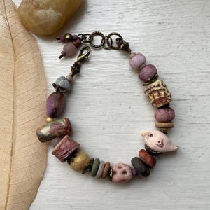 Peut inclure: Un bracelet composé de perles variées dans des tons de rose, violet, vert et doré, incluant une petite breloque en forme d'oiseau en céramique. Les perles sont montées sur un fil métallique avec un fermoir mousqueton et un anneau en bronze.