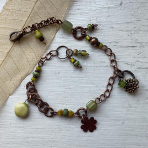 May include: A copper chain bracelet featuring various green beads, including round, cylindrical, and translucent glass shapes. The bracelet includes a copper lobster clasp, a four-leaf clover charm, and a small round textured charm attached to the links.