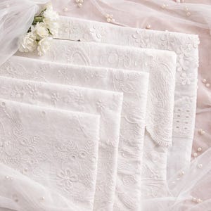 May include: A stack of white fabric squares featuring intricate floral embroidery and eyelet patterns. The edges are finished with delicate scalloped details, and the fabric is layered with sheer white tulle and small white pearl accents.
