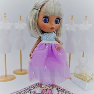May include: A doll with silver hair wearing a light blue and white dress with a lavender tulle skirt and white shoes. The doll is standing on a pink and black patterned rug.
