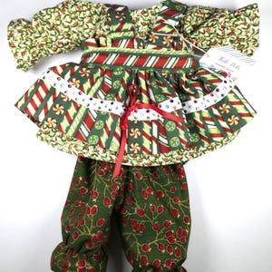 May include: A green and red Christmas-themed doll outfit with a candy cane print skirt, a green top with holly leaves, and green pants with red berries. The outfit has a white ribbon with rhinestones.