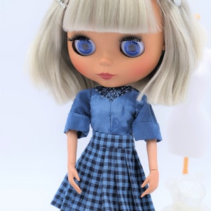 May include: A doll with short, platinum blonde hair, blue eyes, and a blue and white plaid skirt. The doll is wearing a blue shirt with a beaded neckline.