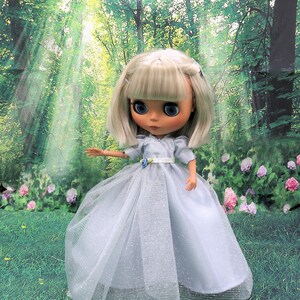 May include: A doll wearing a light blue tulle dress with silver glitter. The doll has blonde hair and blue eyes. The doll is standing in a grassy area with a green forest background.