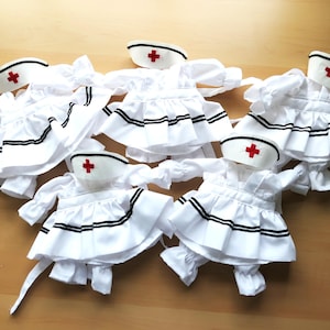 May include: Several white nurse outfits with matching caps are arranged on a wooden surface. Each outfit features a pleated skirt, long sleeves, and black trim. The caps have a red cross emblem.