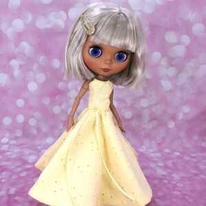 May include: A doll with silver hair wearing a yellow dress with white polka dots. The dress has a white ribbon tied at the waist.