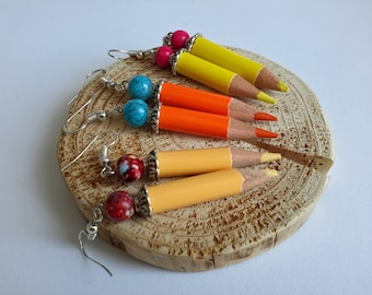 May include: Handmade earrings featuring miniature pencil designs in yellow, orange, and light orange. Each earring has a colorful bead and silver-tone ear wire. The earrings are displayed on a wooden slice.