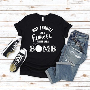 以下が含まれることがあります： 白い文字で「Not Fragile Like a Flower Fragile Like a Bomb」と書かれた黒いTシャツ。Tシャツは白い木製の背景に置かれ、黒いスニーカーと青いジーンズが置かれている。