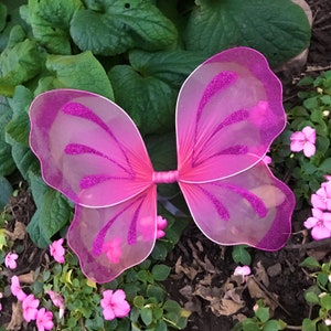 May include: Pink fairy wings with glitter accents. The wings are made of a sheer material with a pink trim and a bow in the center. The wings are designed for children's costumes or dress-up play.