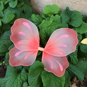 May include: Pink butterfly wings with a gradient from light pink to white. The wings have a delicate, sheer texture and are decorated with glitter.