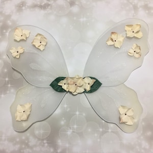 May include: White fairy wings with a delicate, sheer fabric and a row of small, cream-colored flowers across the center. The wings are adorned with additional flowers along the edges.