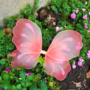 May include: Pink butterfly wings costume accessory with a delicate, sheer fabric and a sparkly design. The wings are spread open and lying on a bed of green plants.