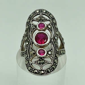 Peut inclure: Une bague en argent avec un motif de filigrane et trois pierres précieuses roses. La bague est posée sur un présentoir transparent.