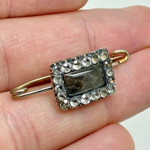 May include: Antique rectangular brooch with a central dark stone, framed by a border of small, clear gemstones. The brooch is set on a gold-toned pin, showcasing a vintage design. A classic piece of jewelry.