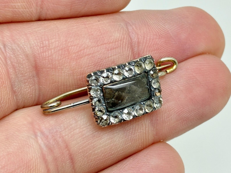 May include: Antique rectangular brooch with a central dark stone, framed by a border of small, clear gemstones. The brooch is set on a gold-toned pin, showcasing a vintage design. A classic piece of jewelry.