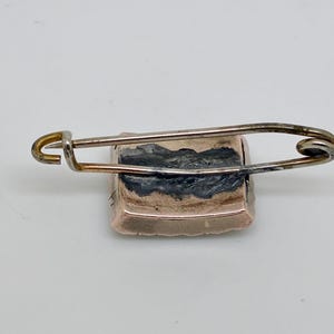 May include: A vintage rectangular brooch with a safety pin clasp. The brooch has a gold-toned frame and a dark, possibly painted, center. The safety pin is silver-toned and attached to the back of the brooch.