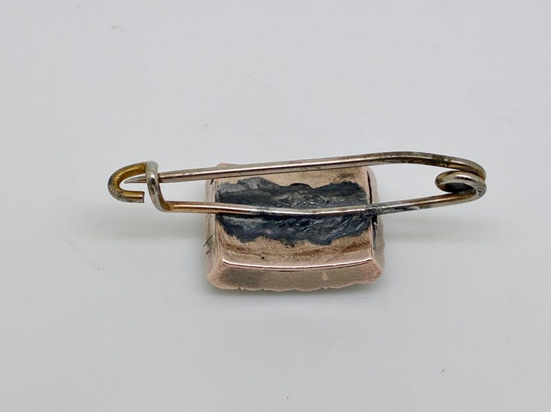 May include: A vintage rectangular brooch with a safety pin clasp. The brooch has a gold-toned frame and a dark, possibly painted, center. The safety pin is silver-toned and attached to the back of the brooch.