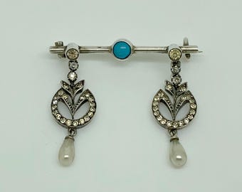 Könnte beinhalten: Eine silberne Brosche mit einem türkisfarbenen Stein in der Mitte und zwei baumelnden Anhängern. Jeder Anhänger zeigt ein florales Design mit einem Perlen-Tropfen.