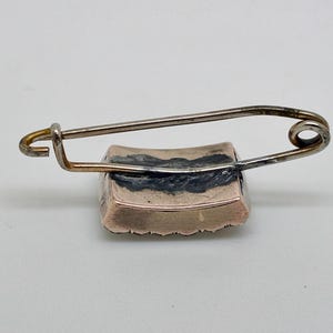 May include: A vintage brooch with a rectangular, gold-toned base and a silver-toned safety pin closure. The brooch has a dark, possibly enameled, design on the top surface. The pin is attached to the top of the brooch.