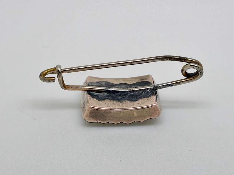 May include: A vintage brooch with a rectangular, gold-toned base and a silver-toned safety pin closure. The brooch has a dark, possibly enameled, design on the top surface. The pin is attached to the top of the brooch.