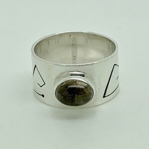 May include: A silver ring with a dark gray stone set in the center. The band of the ring is decorated with geometric designs.