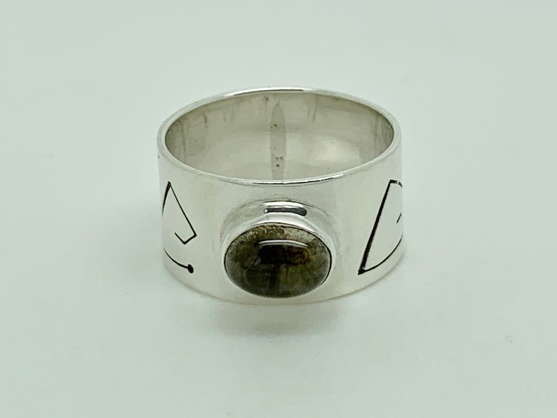 May include: A silver ring with a dark gray stone set in the center. The band of the ring is decorated with geometric designs.