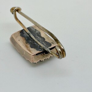 May include: A vintage rectangular brooch with a gold-toned frame and a silver-toned pin. The brooch has a dark, possibly oxidized, center. The pin is attached to the frame with a simple loop.