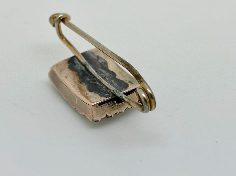 May include: A vintage rectangular brooch with a gold-toned frame and a silver-toned pin. The brooch has a dark, possibly oxidized, center. The pin is attached to the frame with a simple loop.