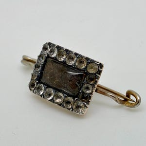 May include: A rectangular silver brooch with a dark center and a border of clear stones. The brooch has a pin backing with a curved end. The design is vintage and elegant.