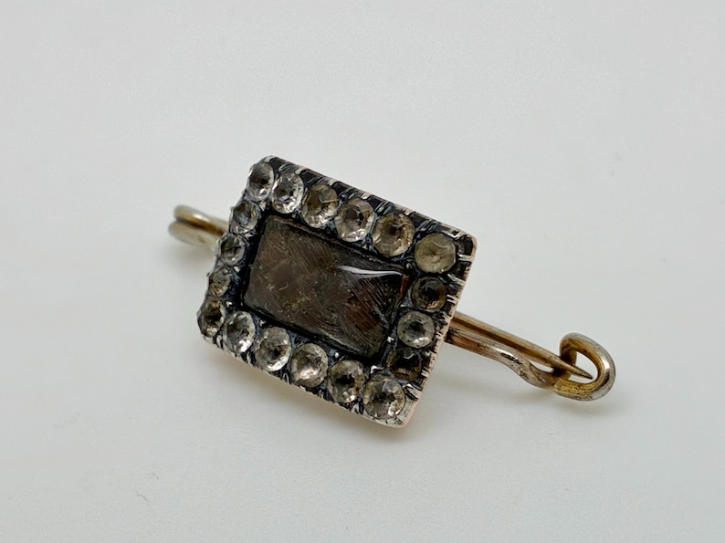 May include: A rectangular silver brooch with a dark center and a border of clear stones. The brooch has a pin backing with a curved end. The design is vintage and elegant.
