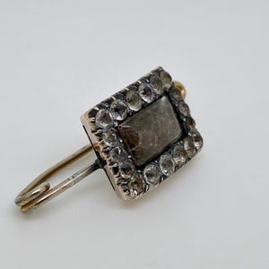 May include: A vintage brooch featuring a rectangular, dark-colored stone at the center, surrounded by a border of smaller, clear stones. The brooch has a gold-colored setting and a silver-colored pin. The overall design is ornate and elegant.