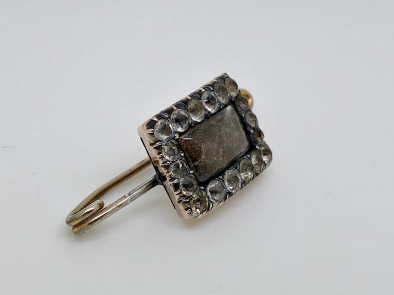 May include: A vintage brooch featuring a rectangular, dark-colored stone at the center, surrounded by a border of smaller, clear stones. The brooch has a gold-colored setting and a silver-colored pin. The overall design is ornate and elegant.