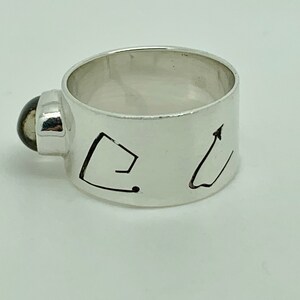 May include: A silver ring with a black stone set into the band. The band is engraved with two abstract designs.