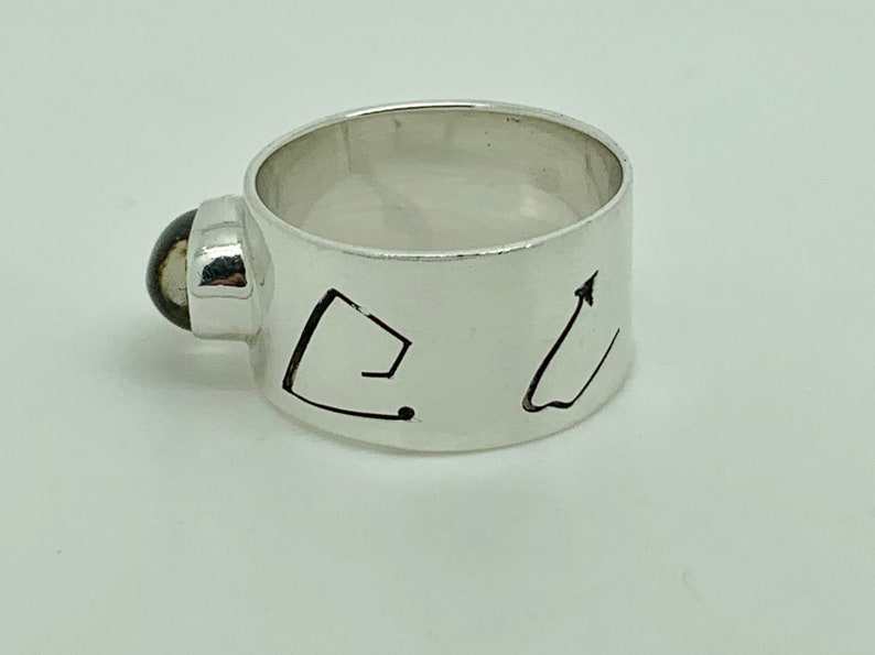 May include: A silver ring with a black stone set into the band. The band is engraved with two abstract designs.