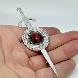 Puede incluir: Un broche de metal plateado con forma de espada, que presenta un medallón central circular con una piedra preciosa de ojo de tigre de color marrón rojizo pulido. El medallón está decorado con patrones en relieve de cardos y hojas.