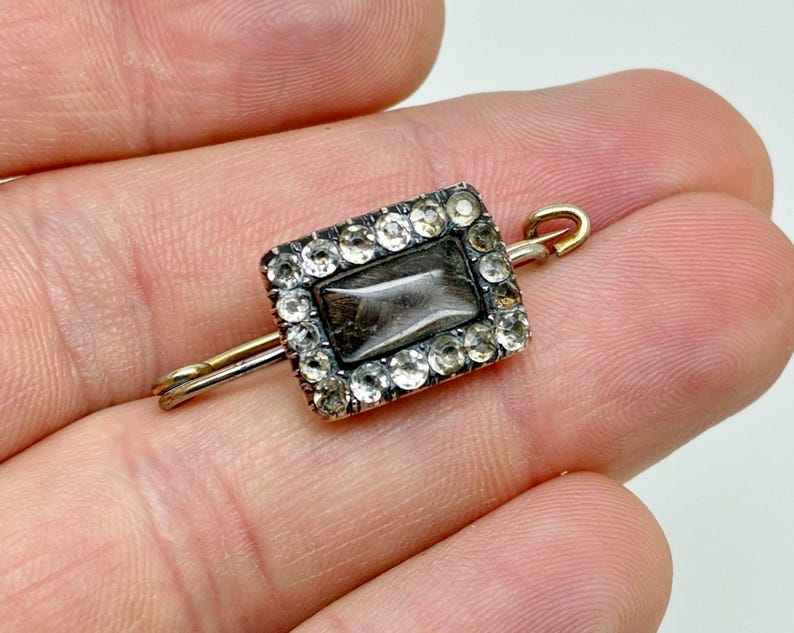 May include: A rectangular antique brooch with a central dark stone, surrounded by clear crystals. The brooch has a gold-toned pin and clasp. The setting is silver-toned, and the piece is held in a hand.