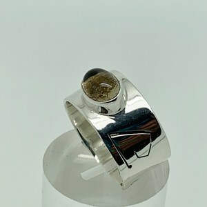 May include: A silver ring with a brown gemstone set in a bezel setting. The ring band is wide and has a geometric design.