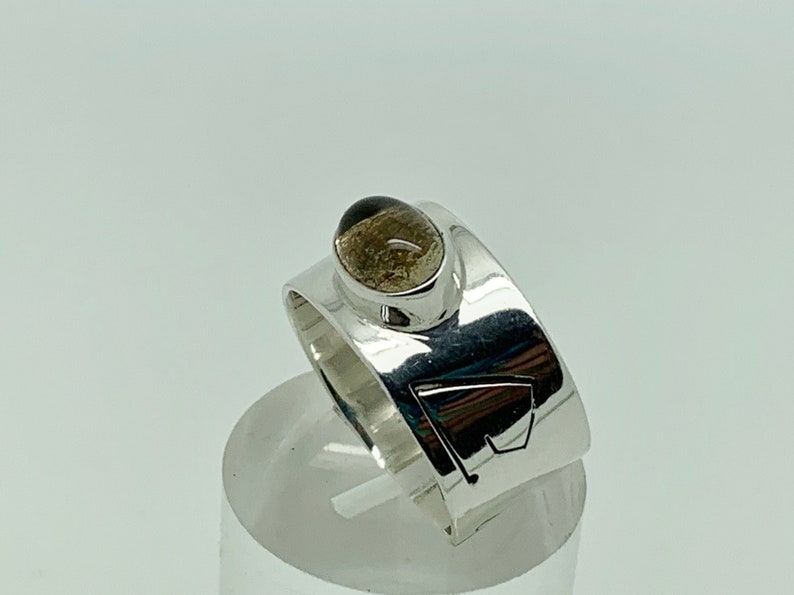 May include: A silver ring with a brown gemstone set in a bezel setting. The ring band is wide and has a geometric design.