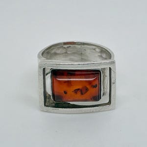 May include: A silver ring featuring a rectangular, polished amber stone set within a geometric, open-frame metal design. The band has a textured, hammered finish, and the amber displays a deep, translucent orange hue with natural inclusions.