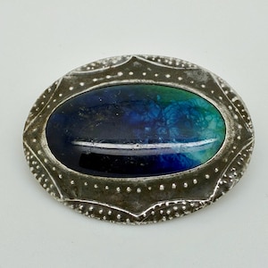 May include: Oval-shaped brooch with a silver-toned frame. The center features a vibrant design with shades of blue and green. The frame has a textured, dotted pattern around the edge, adding a vintage aesthetic.