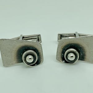 May include: A pair of silver cufflinks with a textured, abstract design. Each cufflink features a small, round ball in the centre.