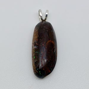 Könnte beinhalten: Ein ovaler Anhänger mit dunkelbrauner Grundfarbe und schillernden grünen und orangenen Einschlüssen. Er hat eine silberfarbene Öse zum Anbringen einer Kette. Der Stein hat ein natürliches, organisches Aussehen.