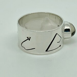 May include: A silver ring with a dark gray gemstone set on the side. The ring has a wide band with two abstract designs engraved on it.
