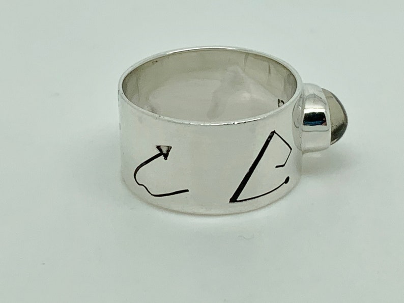 May include: A silver ring with a dark gray gemstone set on the side. The ring has a wide band with two abstract designs engraved on it.